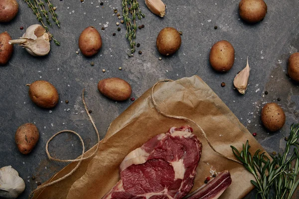 Tendido plano con filete de carne cruda sobre papel de hornear con especias y patatas sobre fondo gris - foto de stock