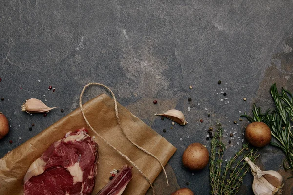 Vue du dessus du steak de viande crue sur papier cuisson avec épices, herbes et pommes de terre sur fond gris — Photo de stock