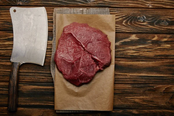 Top view of raw meat on baking paper with butcher knife on wooden background — Stock Photo