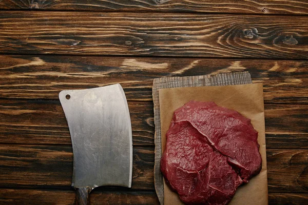 Vista superior de la carne cruda sobre papel de hornear con cuchillo de carnicero sobre fondo de madera - foto de stock