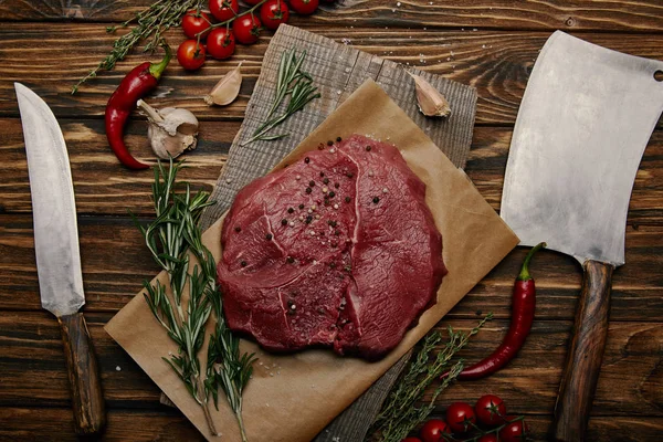 Flat lay with raw meat on baking paper with kitchen knives and vegetables on wooden background — Stock Photo