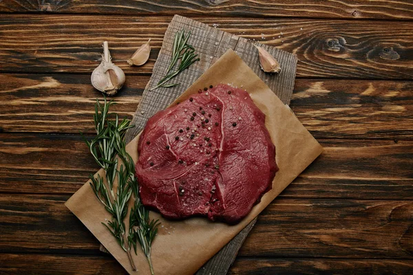 Tendido plano con carne fresca sobre papel de hornear con ajo y romero sobre fondo de madera - foto de stock