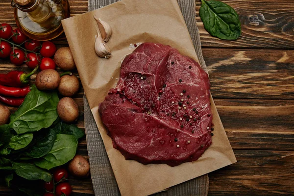 Tendido plano con carne fresca cruda sobre papel de hornear con verduras y espinacas sobre fondo de madera - foto de stock