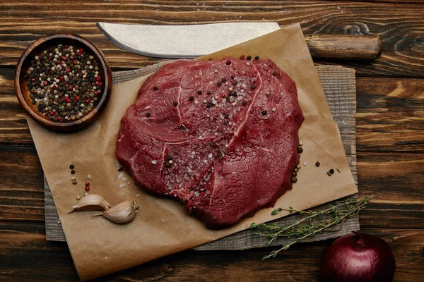 Tendido plano con carne fresca cruda sobre papel de hornear con especias y cuchillo sobre fondo de madera - foto de stock