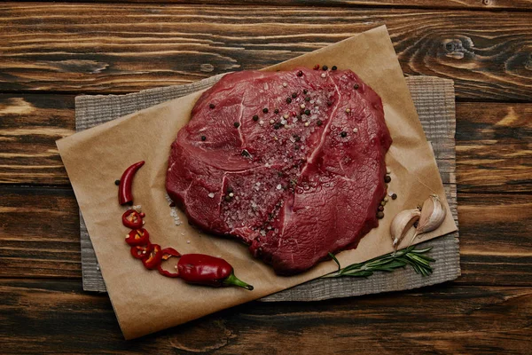 Вид на свежее сырое мясо на бумаге для выпечки с нарезанным перцем чили и чесноком на деревянном фоне — стоковое фото