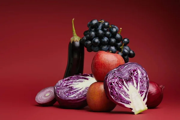 Vista de cerca de manzanas, uvas, berenjenas, repollo y cebollas sobre fondo rojo - foto de stock