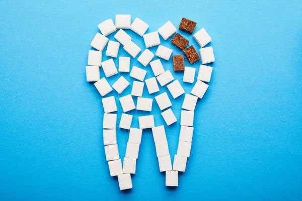 Vista superior de los cubos de azúcar blanco y moreno dispuestos en signo dental sobre fondo azul, concepto de caries dental - foto de stock