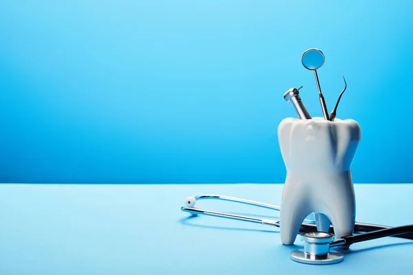 Vista de perto do modelo de dente branco, estetoscópio e instrumentos dentários inoxidáveis no fundo azul — Fotografia de Stock