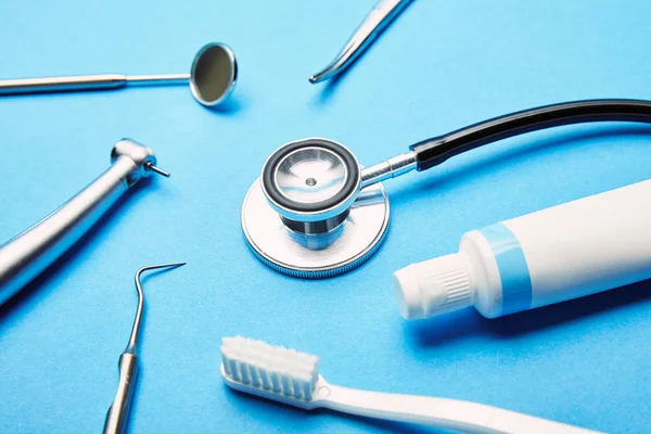 Крупный план расположения стерильных стоматологических инструментов, стетоскопа, зубной щетки и зубной пасты на синем фоне, концепция стоматологической помощи — стоковое фото