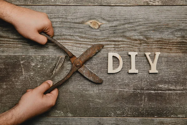 Partial view of man holding vintage rusty carpentry scissors on wooden background with diy sign — Stock Photo