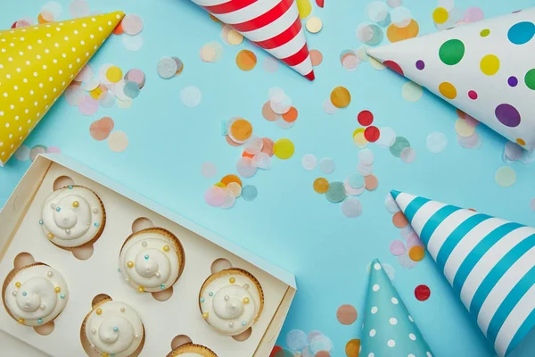 Top view of tasty cupcakes, party hats and colorful confetti on blue background — Stock Photo