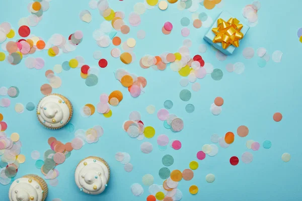 Top view of tasty cupcakes, confetti and gift on blue background — Stock Photo