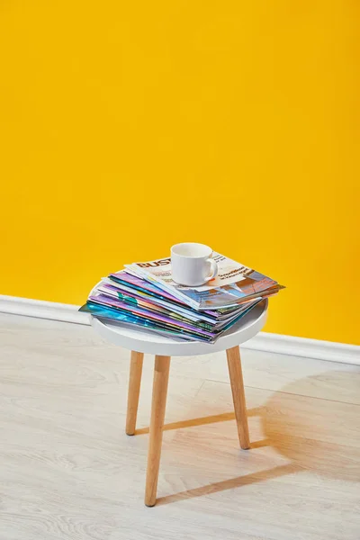 Mesa de café com revistas e copo branco perto da parede amarela — Fotografia de Stock