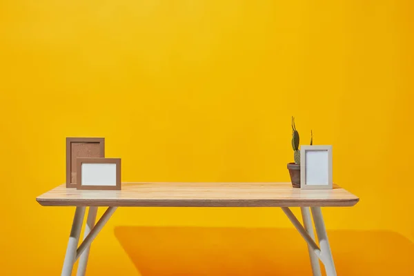 Деревянный стол с пустыми фоторамками и кактусом в цветочном горшке на желтом — стоковое фото