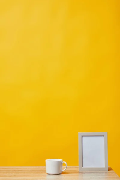 Empty photo frame and cup of coffee at wooden table isolated on yellow — Stock Photo