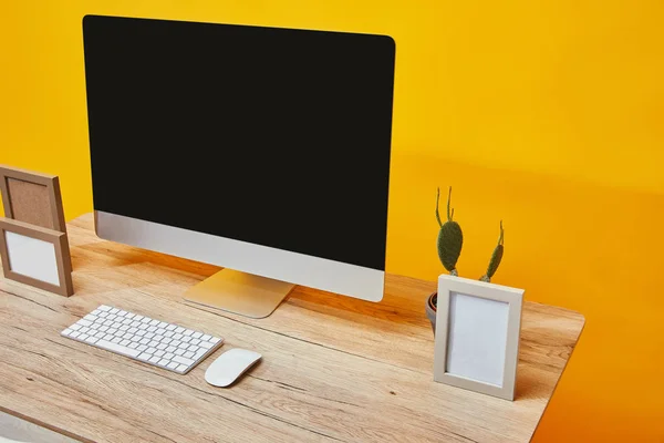 Computador, molduras para fotos e cacto sobre mesa de madeira no fundo da parede amarela — Fotografia de Stock