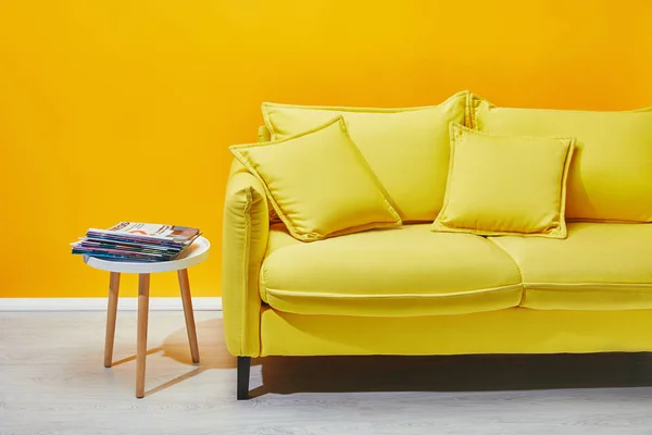 Sofá con almohadas y mesa de centro con prensa cerca de la pared amarilla - foto de stock