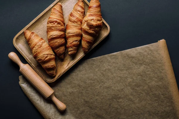 Draufsicht auf Blech mit Croissants, Nudelholz und Backpapier auf schwarzem Tisch — Stockfoto