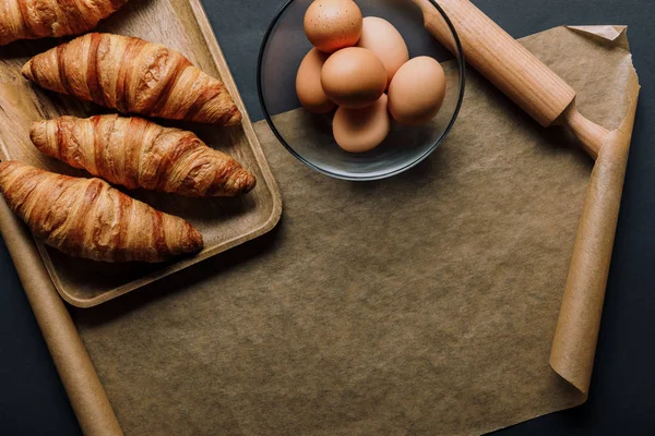 La puesta plana con huevos en un tazón, bandeja con croissants, rodillo y papel de hornear sobre la mesa negra - foto de stock