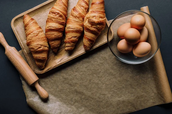 Draufsicht auf Blech mit Croissants, Nudelholz und Backpapier auf schwarzem Tisch — Stockfoto