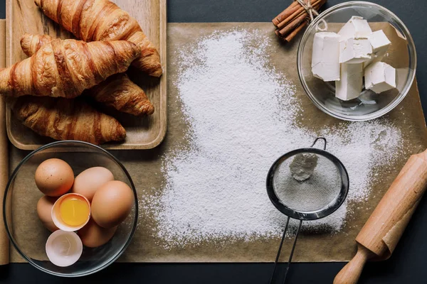Flat lay with baking paper covered by flour, croissants and ingredients on table — Stock Photo