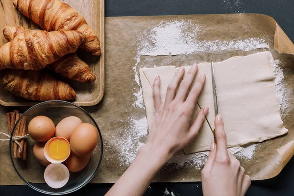 Vue partielle de la pâte à découper femme pour croissants au couteau sur papier sulfurisé recouvert de farine près des ingrédients — Photo de stock