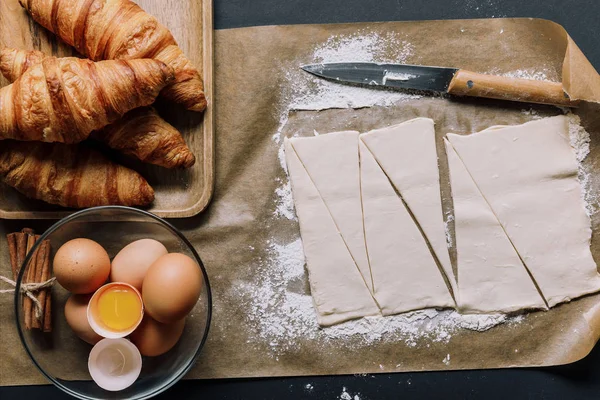 Vista sopraelevata della carta da forno con coltello, pasta per cornetti, uova e cannella in superficie — Foto stock