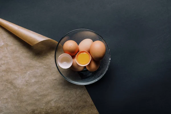 Vista superior de los huevos en un tazón y papel de hornear sobre una mesa negra - foto de stock