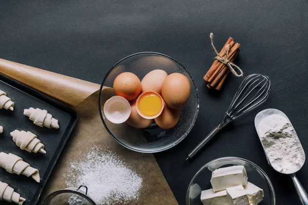 Vista dall'alto degli ingredienti, uova in ciotola e vassoio con pasta per croissant sul tavolo nero — Foto stock