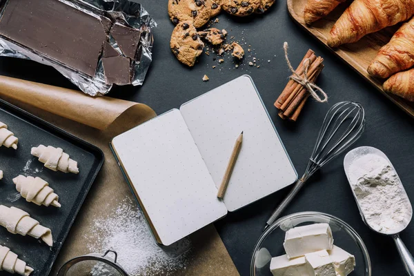 View from above of tray with dough for croissants, ingredients and blank textbook on black table — Stock Photo