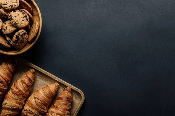 Повышенный вид на поднос с вкусными круассанами и печеньем в миске на черном столе — стоковое фото
