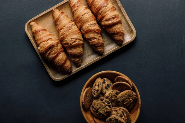 Vista superior de la bandeja con deliciosos croissants y galletas en tazón sobre mesa negra - foto de stock