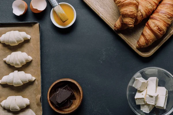 Visão elevada de ingredientes, massa de farinha de croissants na bandeja, gema de ovo com escova na mesa preta — Fotografia de Stock