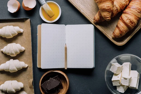 Flache Lage mit leerem Lehrbuch umgeben von Teig für Croissants und Zutaten auf dem Tisch — Stockfoto