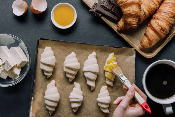 Imagem cortada de mulher escovando massa para croissants na bandeja com papel manteiga perto de ingredientes e xícara de café na mesa — Fotografia de Stock