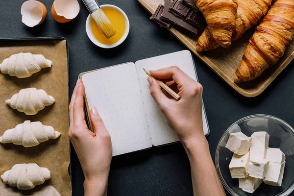 Vista parcial de la mujer escribiendo receta en libro de texto en la mesa con masa para croissants en bandeja e ingredientes - foto de stock