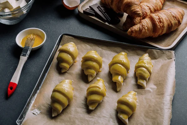 Selektiver Teigfokus für Croissants auf Blech mit Backpapier, umgeben von Zutaten auf schwarzem Tisch — Stockfoto