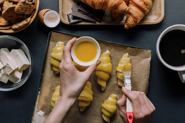 Vista parziale della donna che spazzola la pasta per i croissant su vassoio con carta da forno vicino agli ingredienti e tazza di caffè sul tavolo — Foto stock