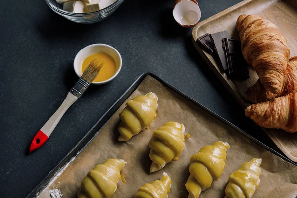 Selektiver Teigfokus für Croissants auf Blech mit Backpapier, umgeben von Zutaten auf schwarzem Tisch — Stockfoto