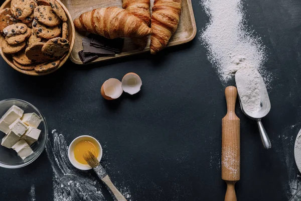 Flache Lage mit Croissants, Keksen und Zutaten auf schwarzem Tisch mit Mehl — Stockfoto