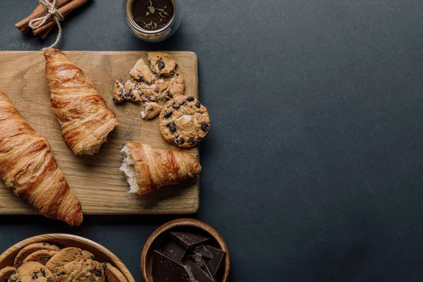 Vista dall'alto della tavola di legno, caffè, cornetti, cioccolato e biscotti sul tavolo nero — Foto stock