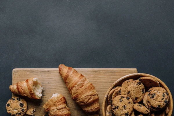 Flat lay with croissants and cookies with chocolate pieces in bowl on black table — Stock Photo