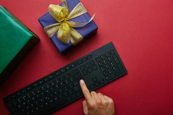 Cropped view of man pushing button on computer keyboard near wrapped boxes on red background — Stock Photo