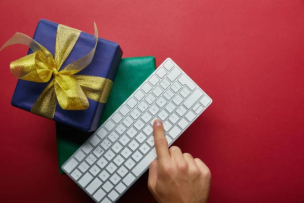 Vue du dessus de l'homme appuyant sur le bouton sur le clavier de l'ordinateur près de cadeaux enveloppés sur fond rouge — Photo de stock