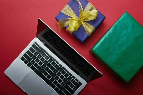 Vista recortada de la computadora portátil cerca de regalos envueltos en fondo rojo — Stock Photo