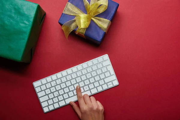 Vue recadrée de la femme appuyant sur le bouton sur le clavier d'ordinateur blanc près des cadeaux sur fond rouge — Photo de stock