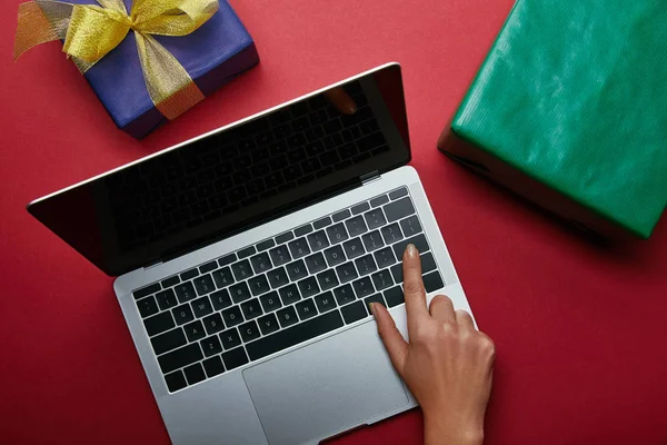 Vista superior de la mujer pulsando el botón en el teclado del ordenador portátil cerca de los regalos en fondo rojo — Stock Photo