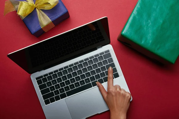 Vista superior de la mujer pulsando el botón en el teclado del ordenador portátil cerca de regalos en fondo rojo — Stock Photo