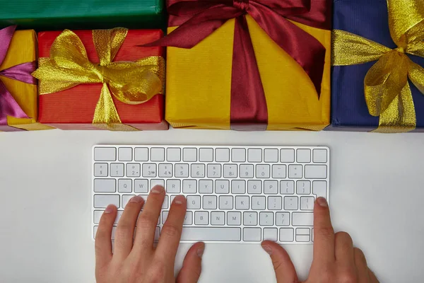 Вид зверху людини, що друкує на клавіатурі комп'ютера біля загорнутих подарунків на білому тлі — стокове фото
