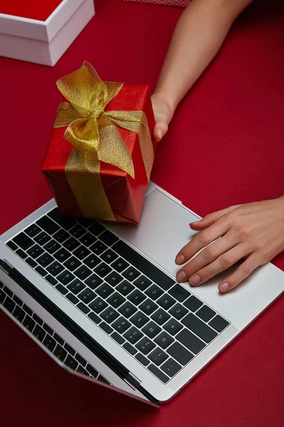 Cropped view of woman holding wrapped gift near laptop on red background — Stock Photo